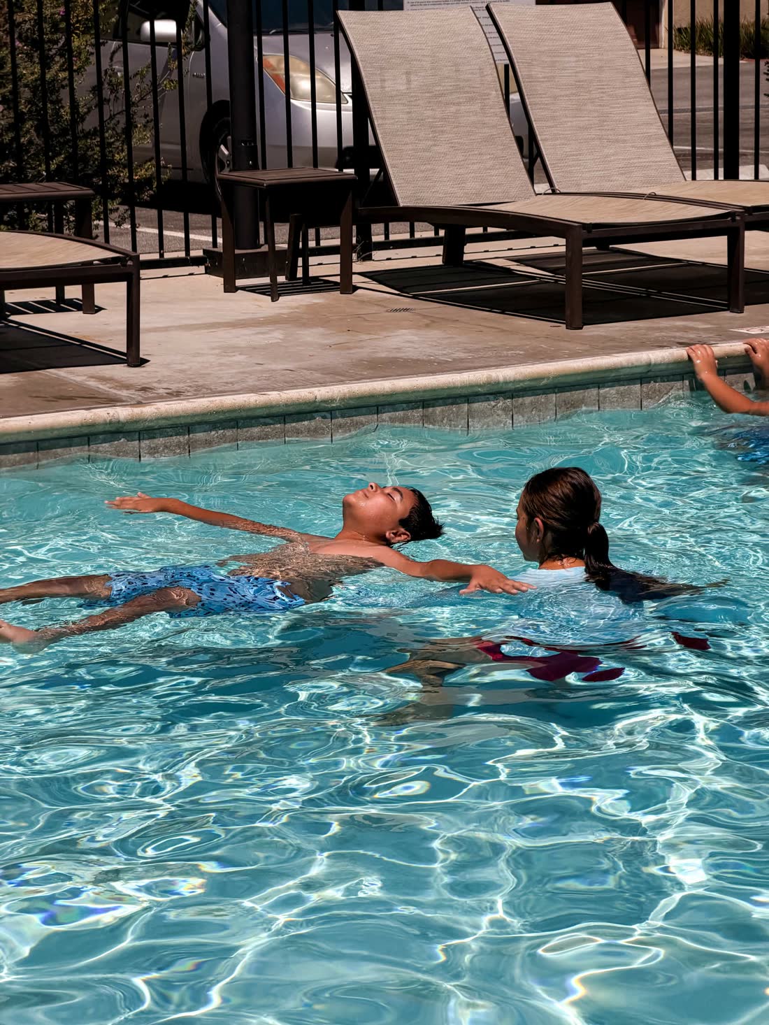 A young swimmer holding a back float at the surface while Emmy supports him from beside.