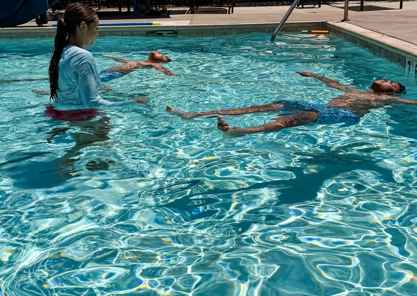 Two swimmers holding independent back floats while Emmy watches from standing depth.